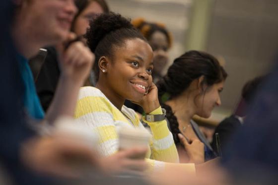 一个穿着黄色毛衣的女人坐在演讲厅里的照片, 上课时微笑并记笔记. 