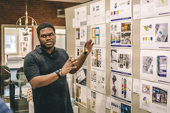 图为一名波胆网站设计专业的学生在一面墙前展示印刷设计材料. 